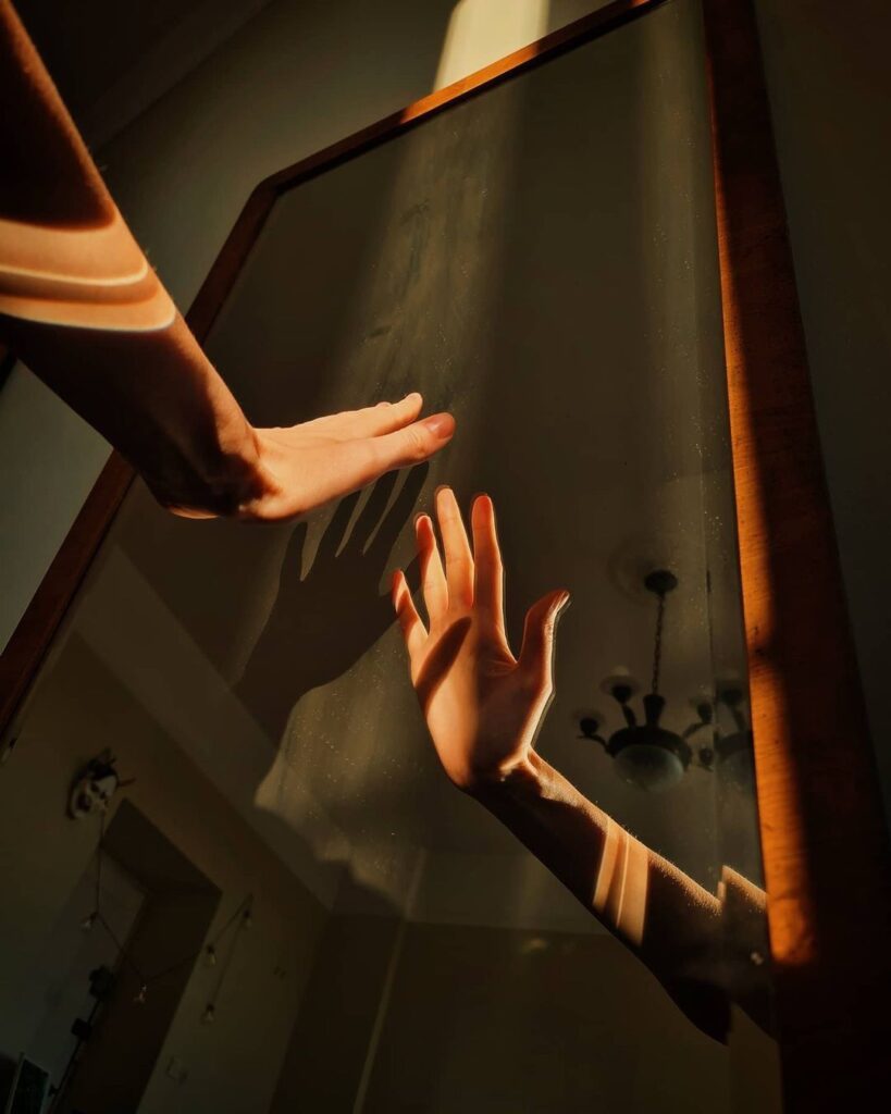Mão feminina alcançando o reflexo no espelho, em um ambiente com iluminação quente e nostálgica. A foto ilustra a sensação de perda de identidade, o desconhecimento de si mesma e o processo de autoconhecimento na psicoterapia