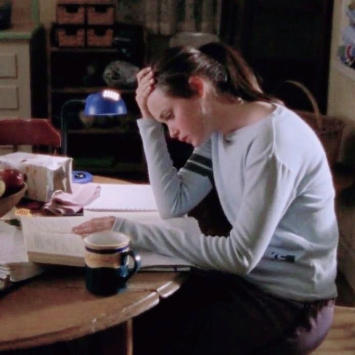 Fotografia da personagem Rory Gilmore, da série Gilmore Girls, sentada à mesa de estudos à noite. Ela veste um suéter cinza claro, tem os cabelos presos e apoia a cabeça com a mão esquerda, demonstrando exaustão e cansaço. Em primeiro plano, sobre a mesa de madeira, há um livro aberto e uma caneca de café